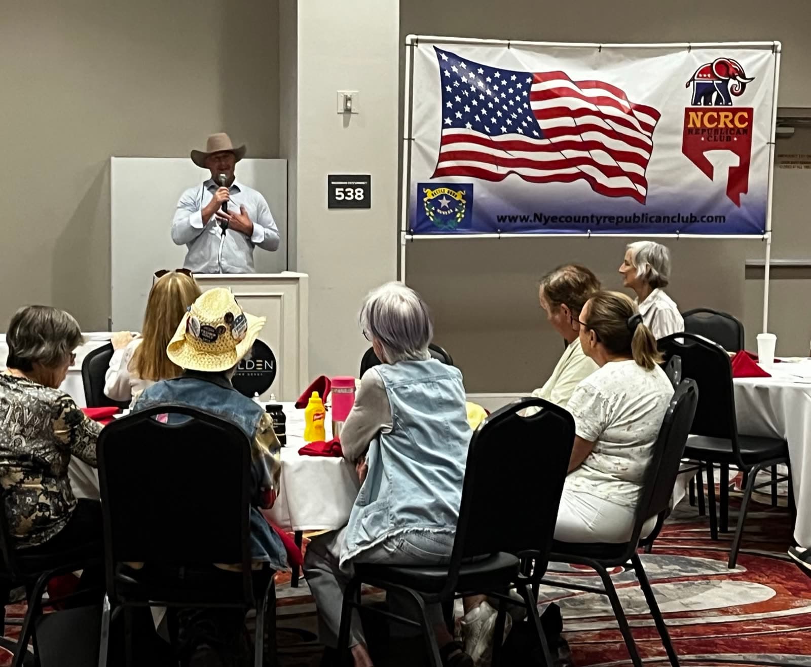 A club luncheon at the Pahrump Nugget: a speaker at the microphone beside the club's flag banner, with members seated at round tables listening.